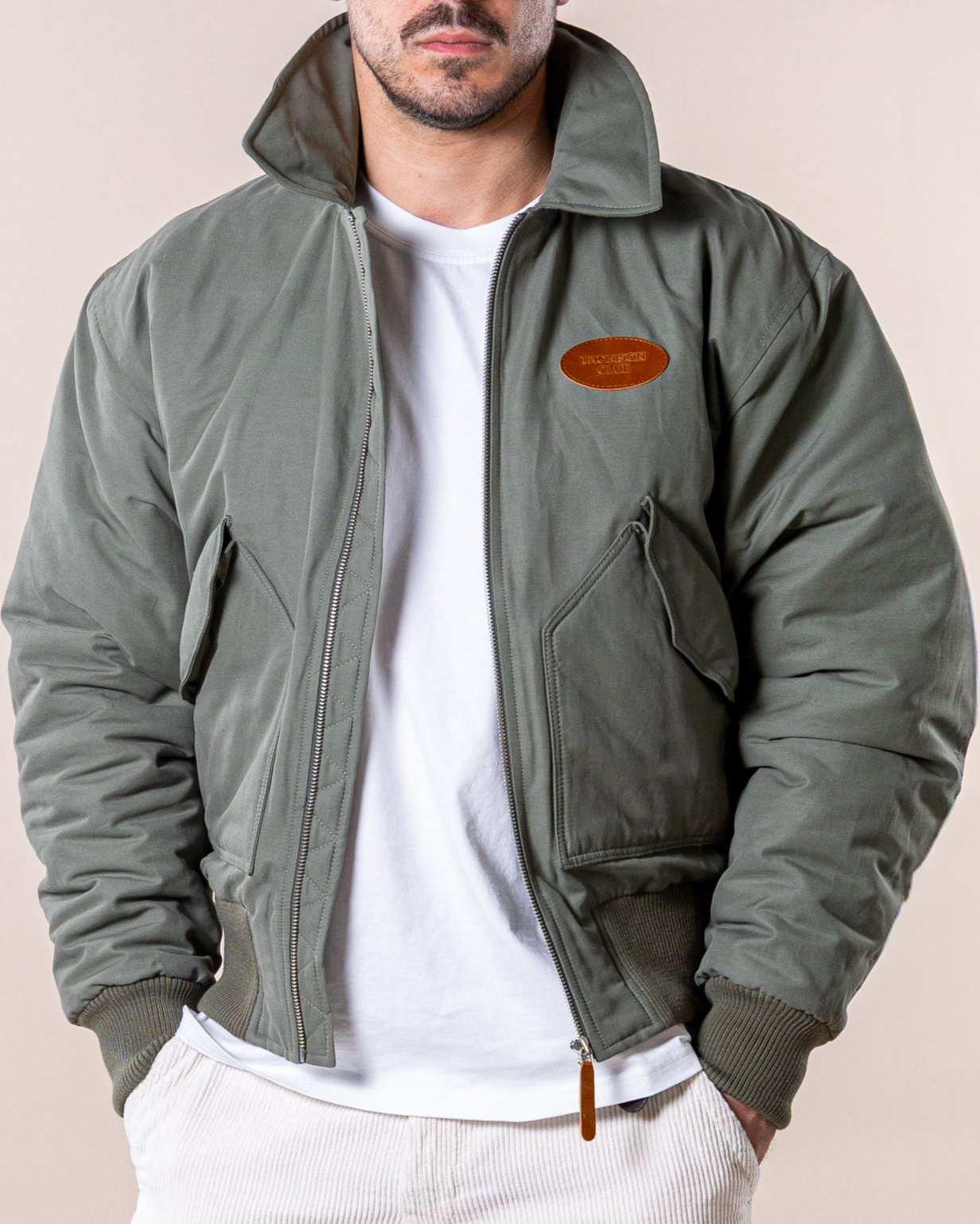 Casaco bomber de homem caqui, feito de tecido esmerilado premium (60% algodão, 40% poliéster). Acolchoado para máximo conforto, com corte regular, gola clássica e punhos/bainha em rib 2X2 para ajuste flexível. Inclui bolso interno, patch de couro com logotipo Thomson Club e puxador TC personalizado de couro. Design icônico e atemporal, perfeito para atitude e confiança.