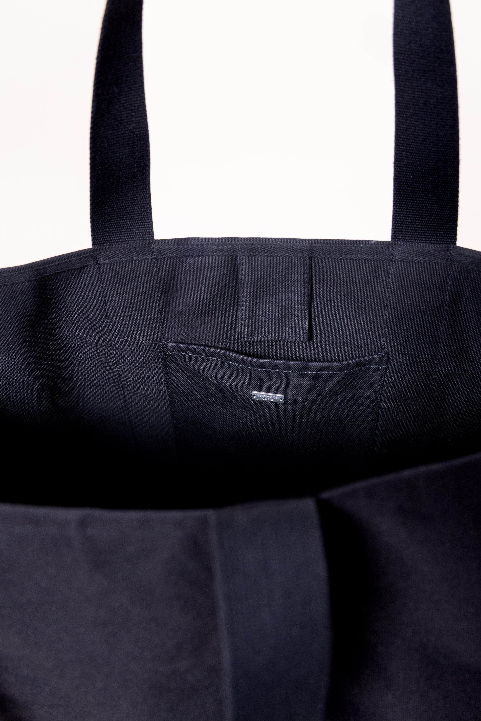 Saco estruturado e espaçoso, ideal para o dia a dia. Feito em lona pesada 500g/m², 100% algodão preto, combina resistência e estilo. Com alças reforçadas para uso na mão ou ombro, mede 38x18x36 cm. Possui bolso interno com tag metálica prateada, logotipo Thomson Club bordado, etiqueta bandeira TC e botão de pressão. Qualidade e design atemporal.