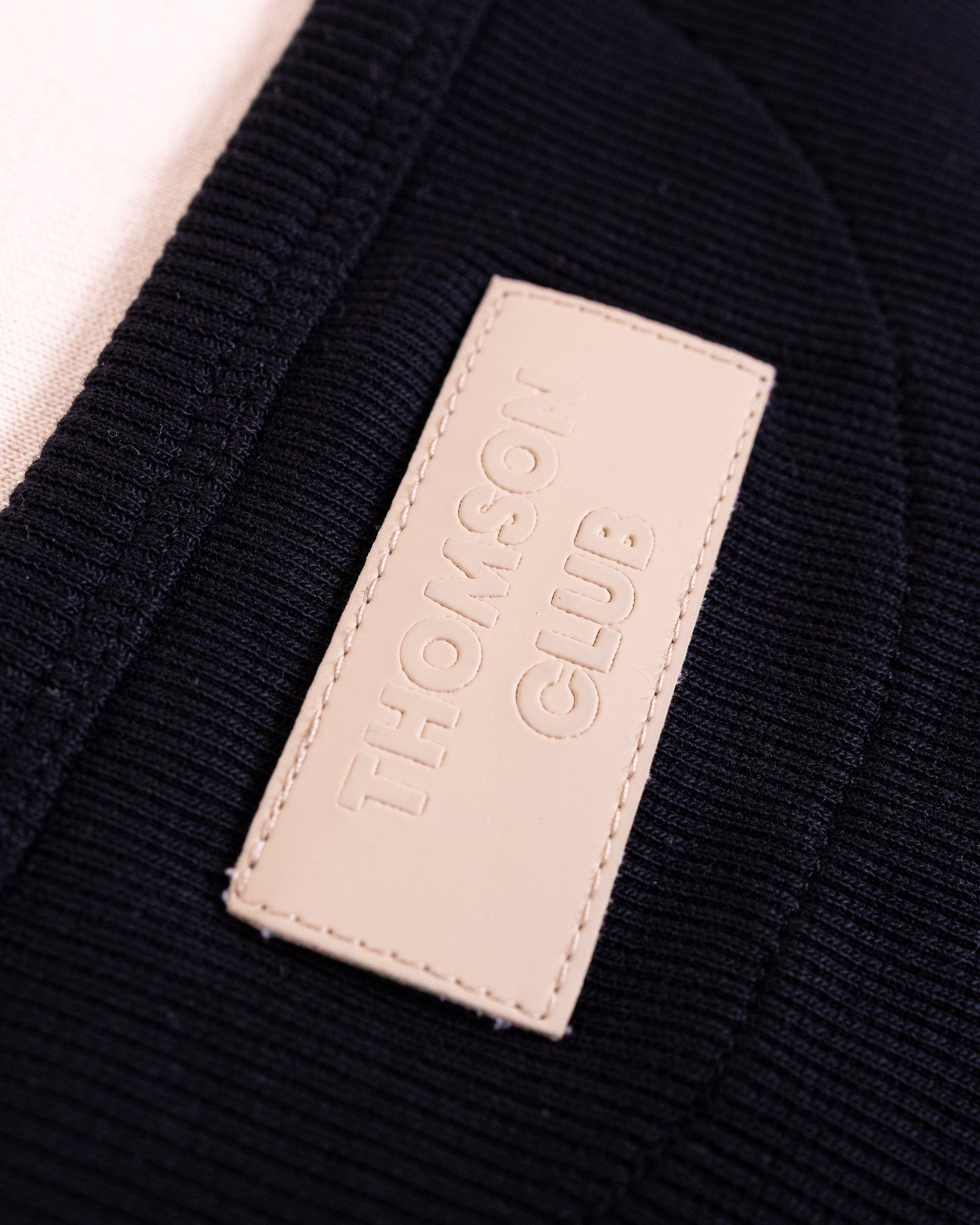 Detalhe do patch em tom Ivory com logo Thomson Club em relevo positivo no vestido preto